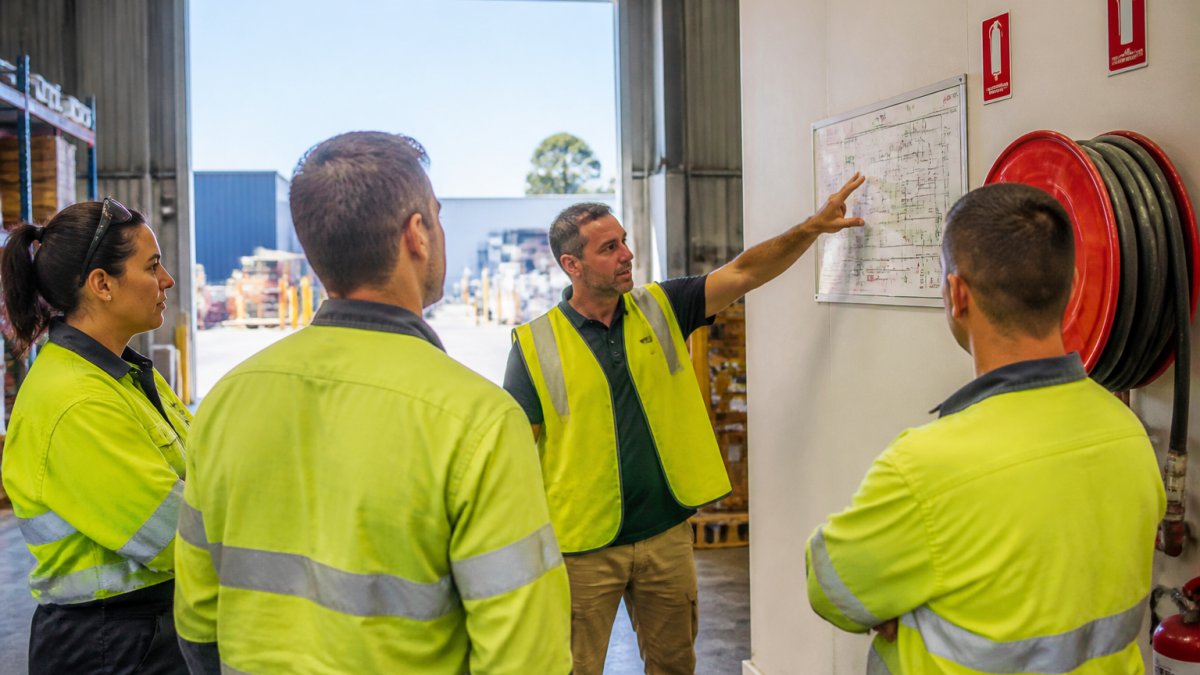 A small group of employees gathered around a fire warden in a warehouse space, pointing at a fire evacuation map on the wall during a toolbox talk, natural daylight through roller doors
