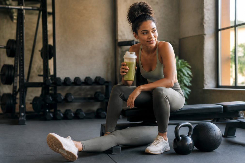 Plant-based athlete in workout gear sitting on a bench with a water bottle and recovery smoothie, stretching legs after intense training session, natural gym lighting