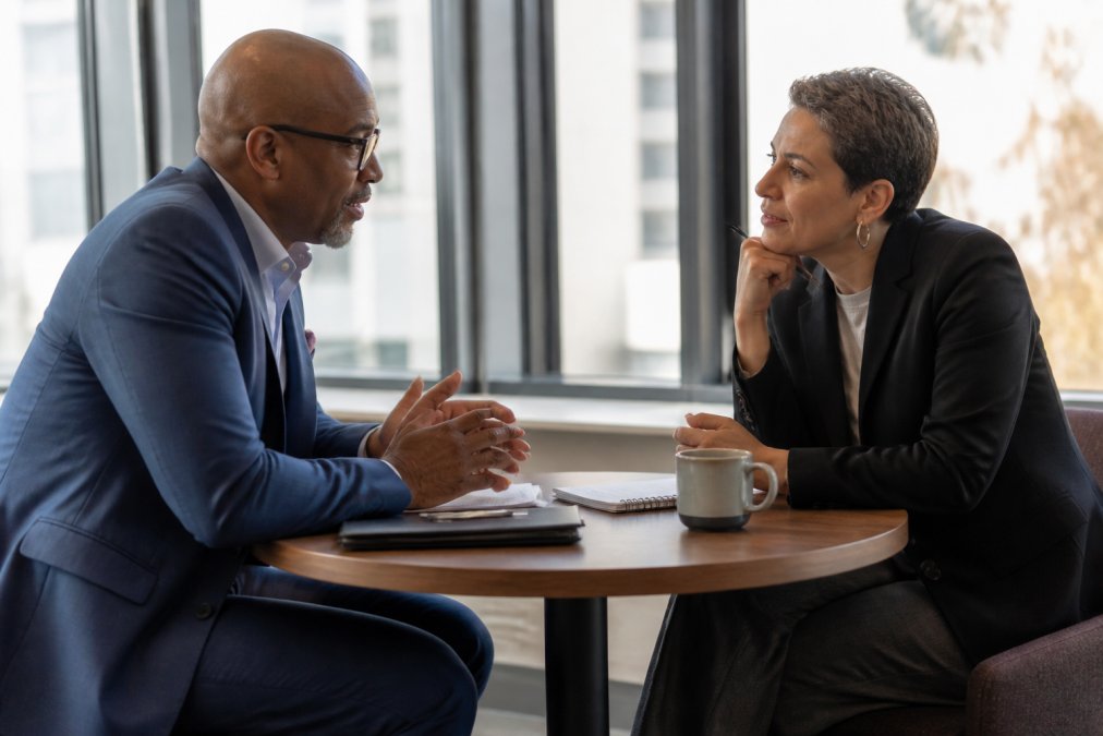 A leadership coach and an executive seated across from each other at a small table in a bright office, notes and a coffee cup between them, both leaning in during a candid conversation
