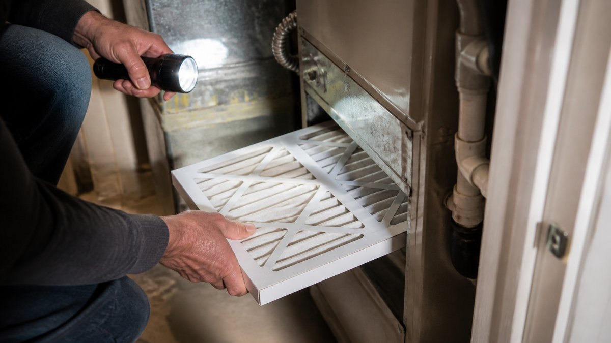Close-up of a homeowner's hands sliding a clean pleated furnace filter into the return slot of a residential utility closet, flashlight glow on dusty ductwork