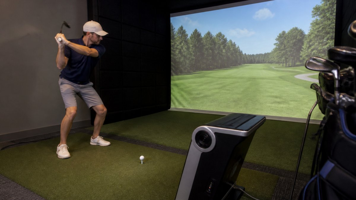 A golfer mid-swing inside an indoor simulator bay, with a projected fairway on the screen and a launch monitor showing ball flight data in the foreground