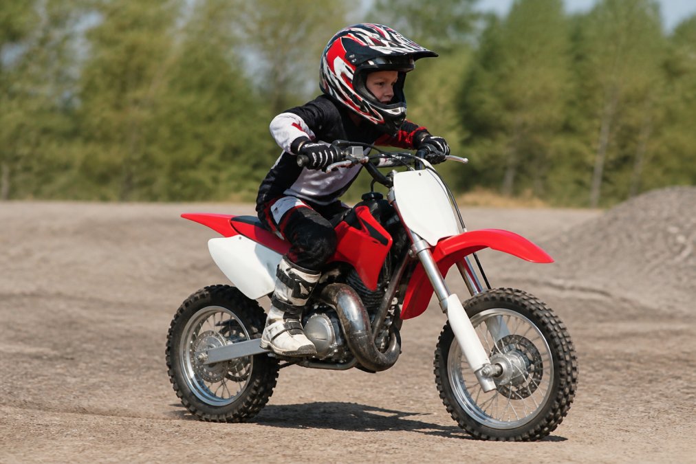 Young rider on a 50cc dirt bike demonstrating proper body positioning with knees gripping the tank, elbows up and out, and head positioned forward over the handlebars, on a flat practice area with natural outdoor lighting
