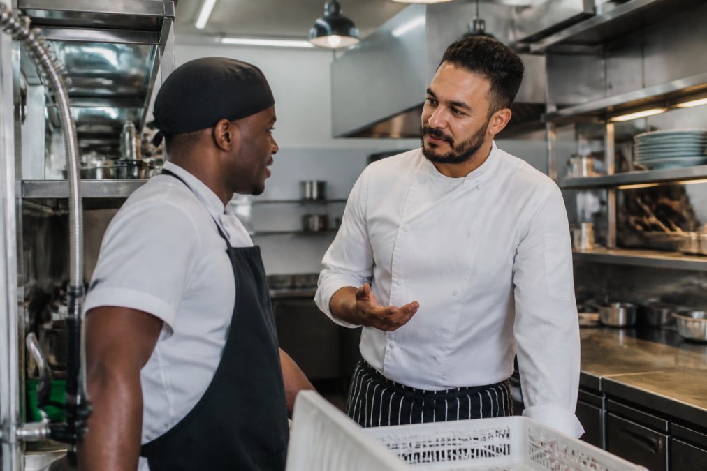 A head chef speaking calmly with a junior kitchen porter in a clean, well-lit commercial kitchen during a quiet moment, both looking relaxed and engaged in conversation