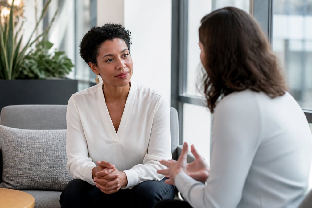 Two managers having a one-to-one conversation in modern office setting with large windows, one listening attentively while the other speaks, conveying trust and open communication, warm natural lighting