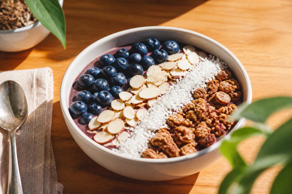 Close-up of a vibrant smoothie bowl topped with fresh blueberries, sliced almonds, shredded coconut, and granola on a bright wooden table with natural morning light