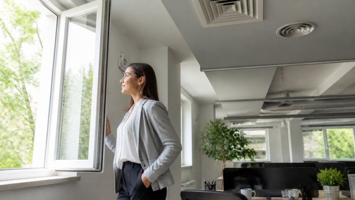 Professional office worker positioned near an open window with natural sunlight streaming in, showing modern workspace with visible ceiling air vents and clean HVAC returns