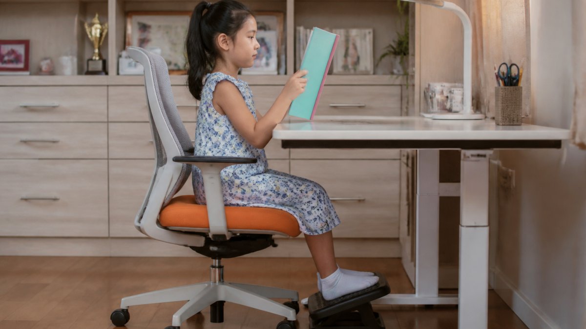 Child sitting upright at desk with proper posture, book positioned at eye level, feet flat on footrest, showing correct 90-90-90 angle positioning with natural office lighting