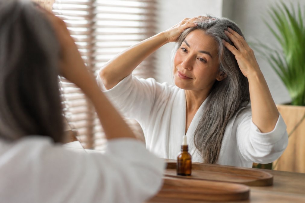 A woman in her 50s with silver-streaked hair sitting at a well-lit vanity, gently massaging oil into her scalp with her fingertips, a small amber bottle of hair oil visible on the counter beside her