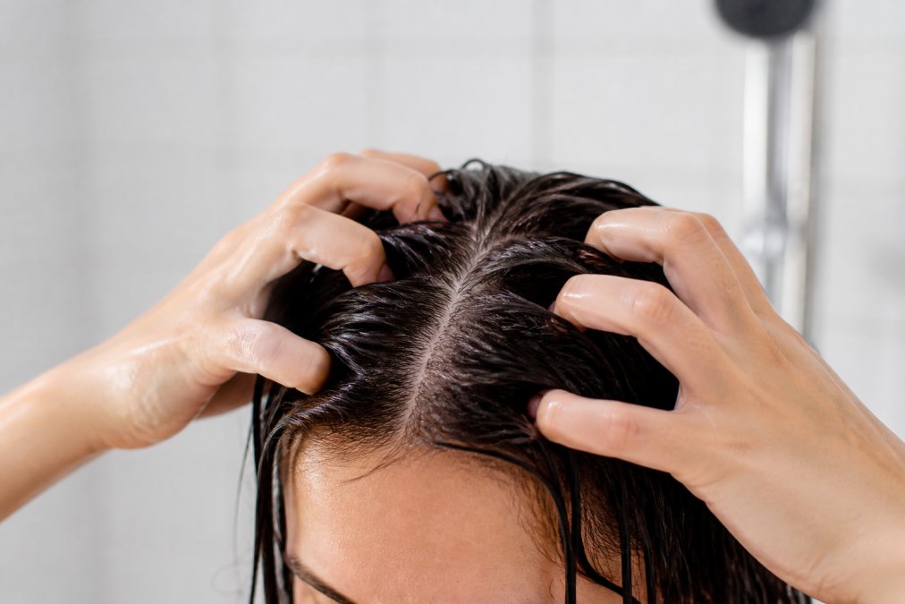 Close-up of a person's hands gently massaging their own scalp with fingertips in a bright bathroom, showing wet hair and a visible scalp parting