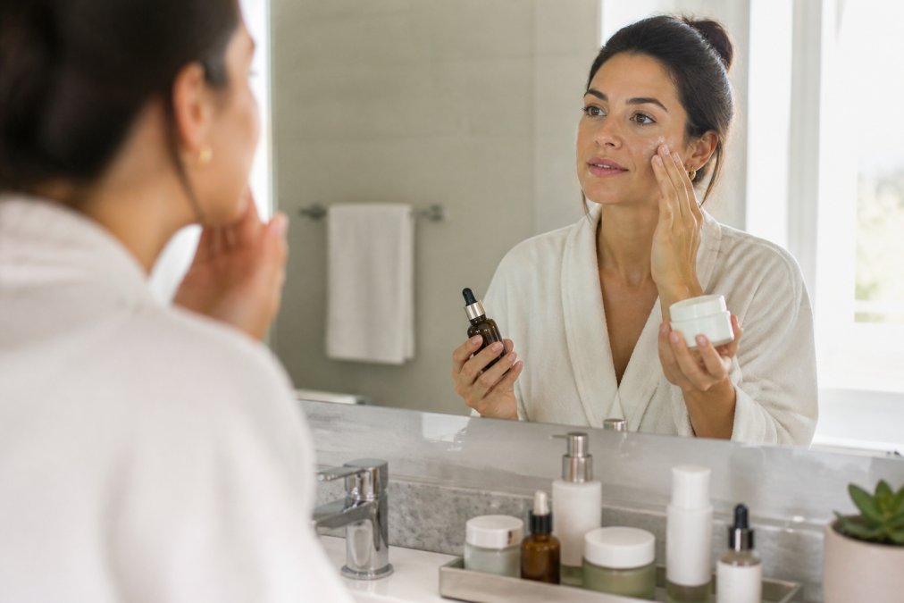 Woman in her 30s applying skincare products at a bright bathroom mirror, holding a retinol serum bottle and moisturizer, with organized skincare bottles on the counter