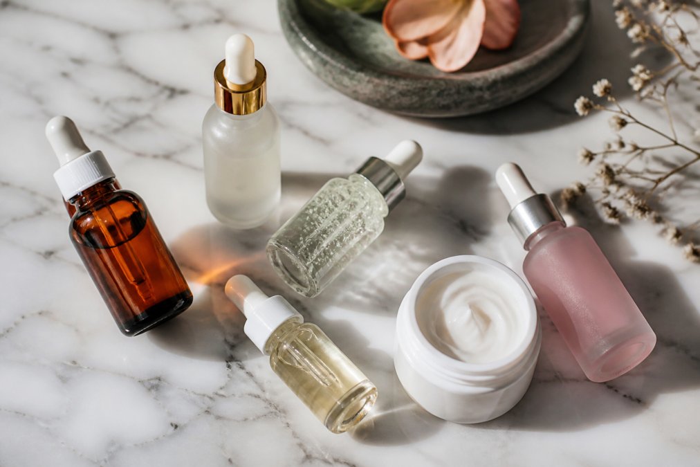 Flat lay of several skincare serums and moisturizer bottles with dropper applicators arranged on a white marble surface, with small ingredient labels visible in soft natural light