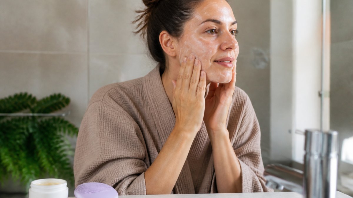 A woman in her thirties at a bathroom sink applying a gentle cleanser to damp skin, with a sonic silicone cleansing device and a repair balm visible on the counter beside her