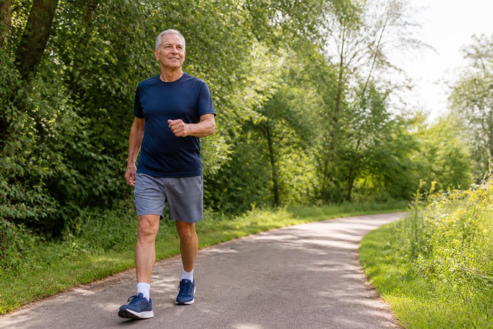 Active man in his 60s performing brisk walking outdoors on a sunny path, wearing comfortable athletic wear and demonstrating good posture with natural lighting