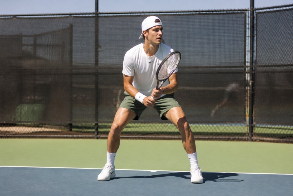 Competitive tennis player at the baseline executing a split step with knees bent, weight balanced on the balls of the feet, shoulders squared, ready to move laterally in bright outdoor court lighting