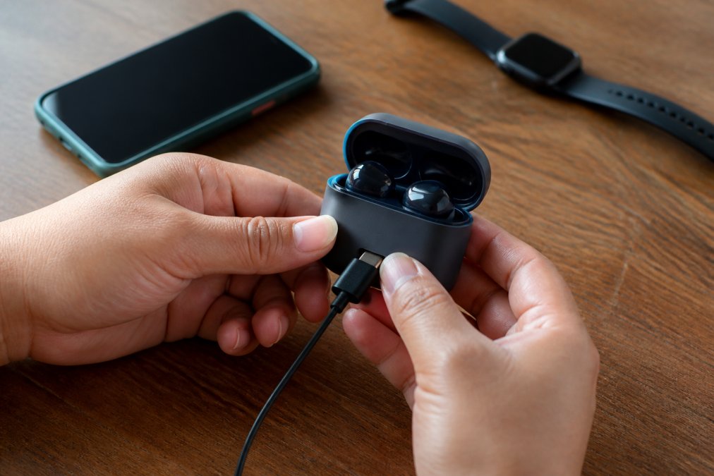 A close-up of hands plugging a USB-C cable into wireless earbuds resting in their charging case on a wooden desk, with a smartphone showing a battery percentage in the background