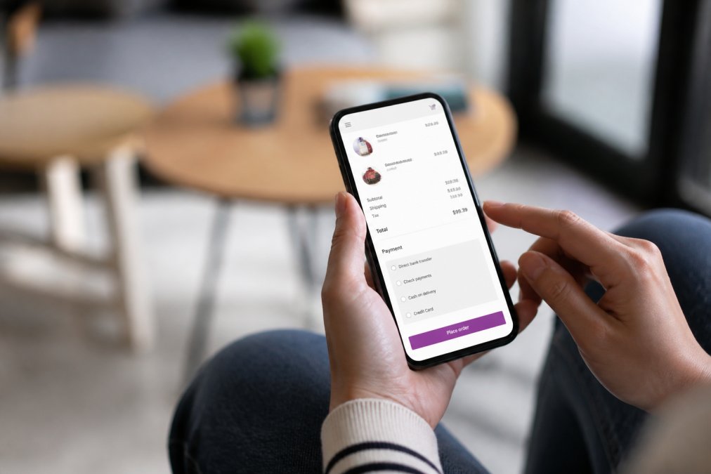 Person's hands holding a smartphone while shopping on a WooCommerce store, with a mobile-optimised checkout page visible on the screen in a bright, modern setting