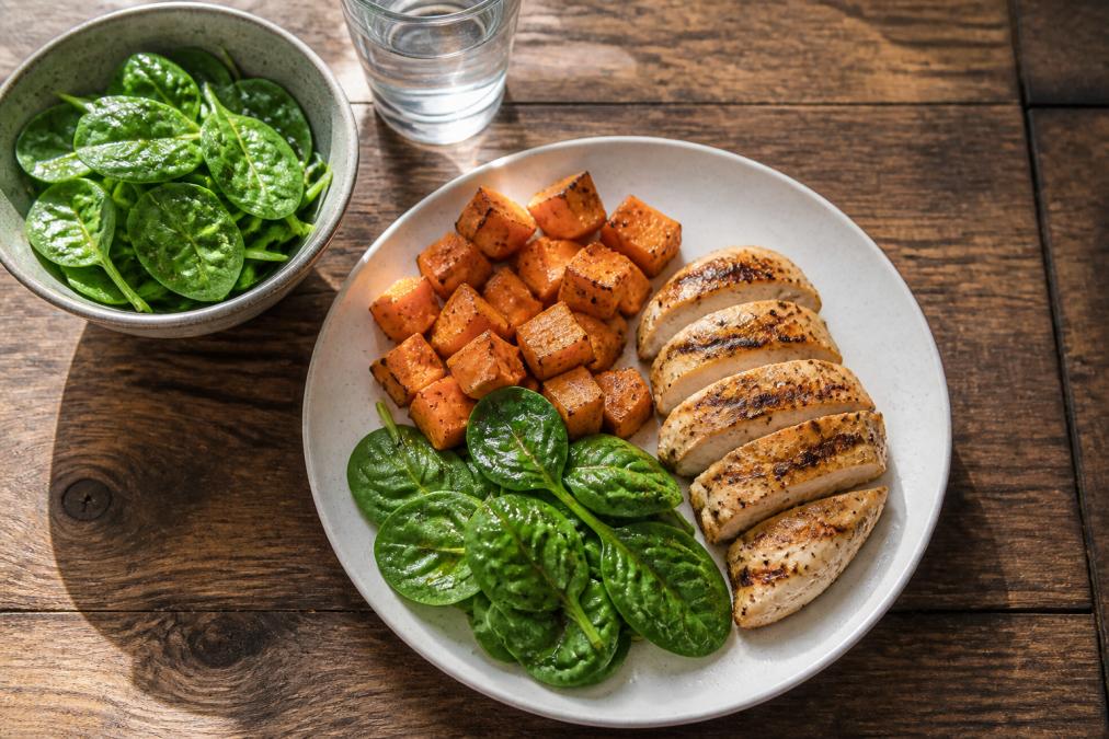 Overhead shot of a recovery meal spread with grilled chicken breast, roasted sweet potato, fresh spinach salad with olive oil, and a glass of water on a wooden table in natural daylight