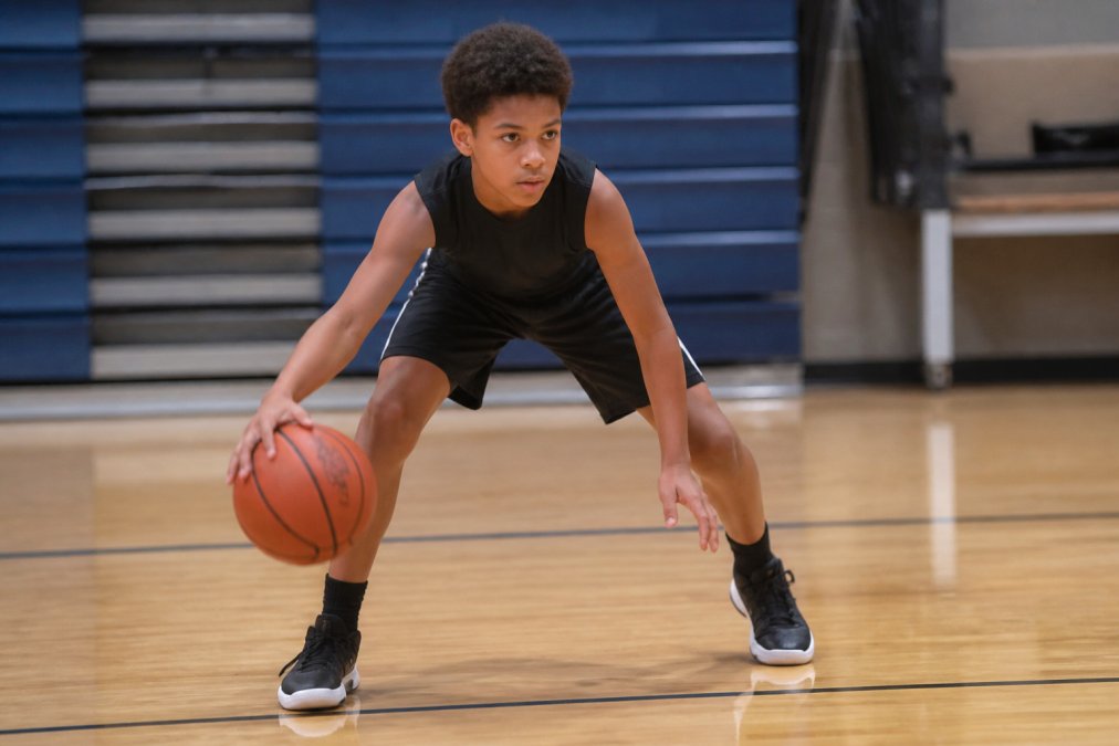 Young basketball player in athletic stance with deep knee bend, dribbling the ball at knee height with proper finger pad contact and focused expression during practice