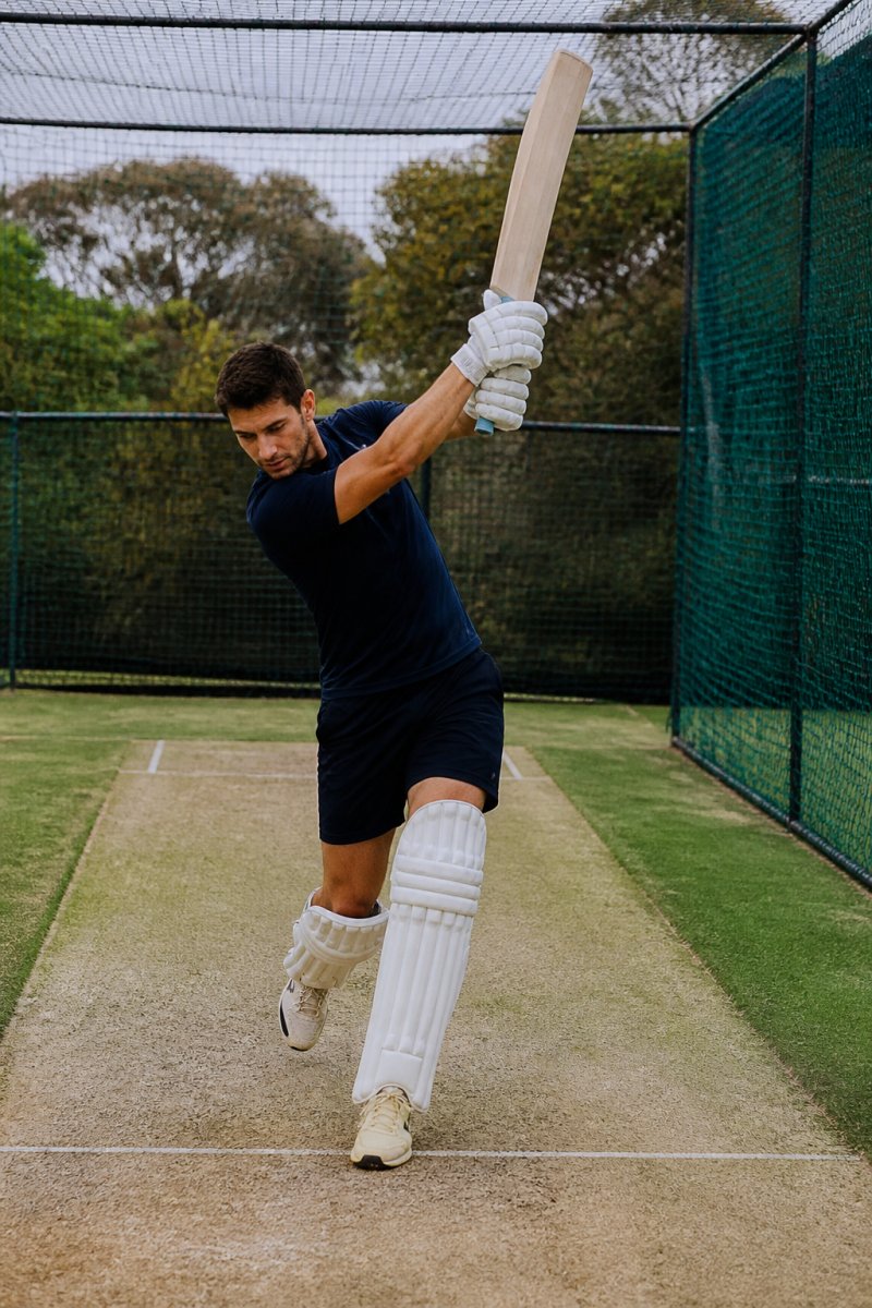 A club cricketer in training gear performing shadow batting drills in an outdoor practice net, mid-swing with a cricket bat, focus on footwork and bat path, natural daylight