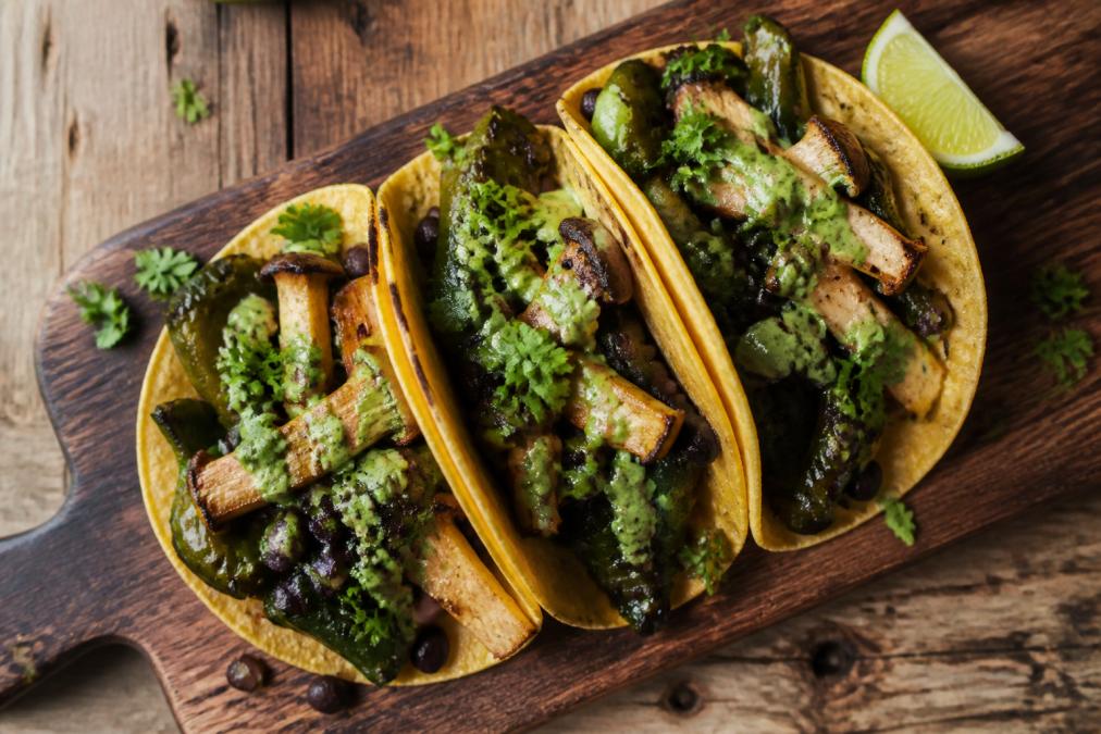 Overhead shot of finished vegetarian tacos with charred poblano peppers, king oyster mushroom pieces, black beans, fresh cilantro garnish, lime wedge, and drizzle of aji verde sauce on warm corn tortillas arranged on a rustic wooden board with natural lighting