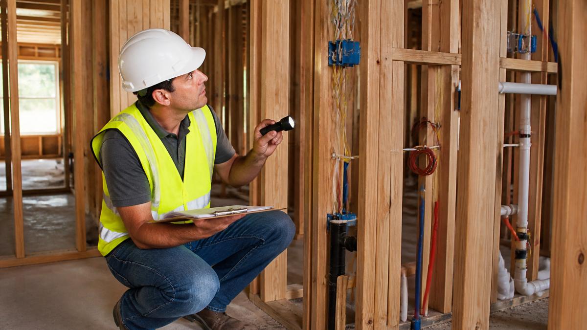 A home inspector in a hard hat and safety vest examining exposed wood framing, electrical wiring, and plumbing rough-ins inside a house under construction, with a clipboard and flashlight in hand