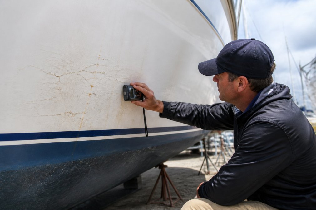 Marine surveyor using a moisture meter on a yacht hull, examining visible gelcoat cracks and discoloration in bright natural light