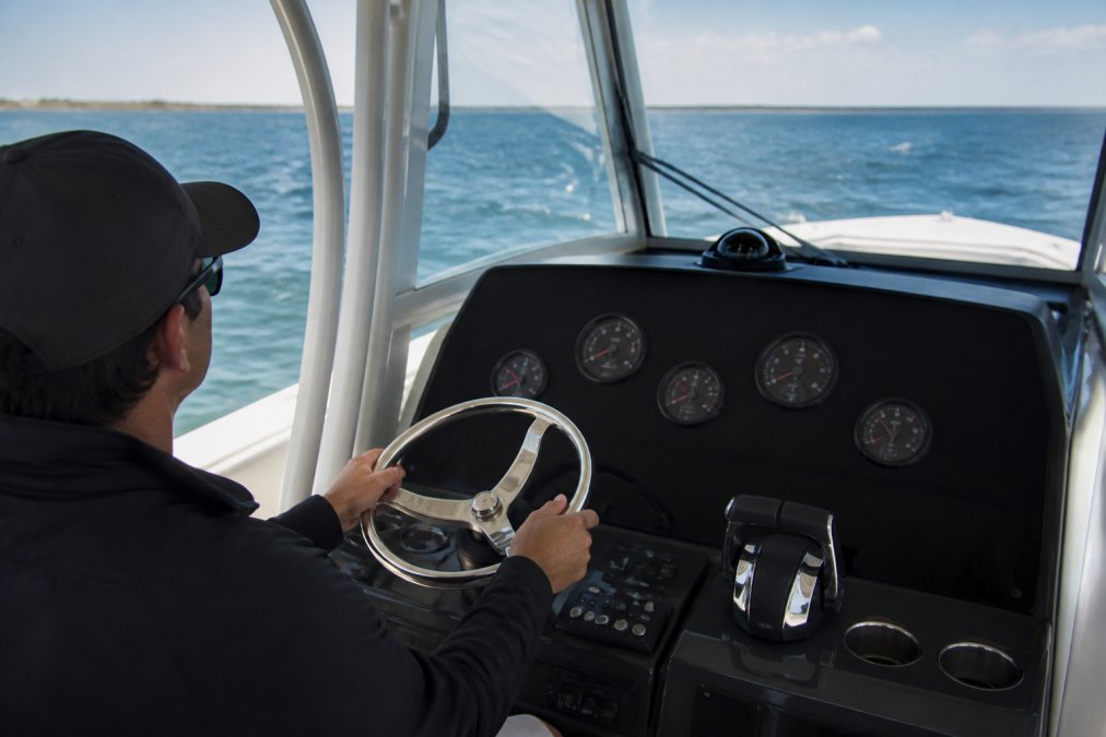 Captain at the helm of a center console yacht during sea trial, hands on the wheel with water visible through the windscreen and engine gauges visible on the dashboard
