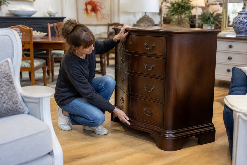 A person kneeling beside a wooden dresser in a bright consignment shop, gently pushing the top corner to test for wobbles while examining the base for water stains