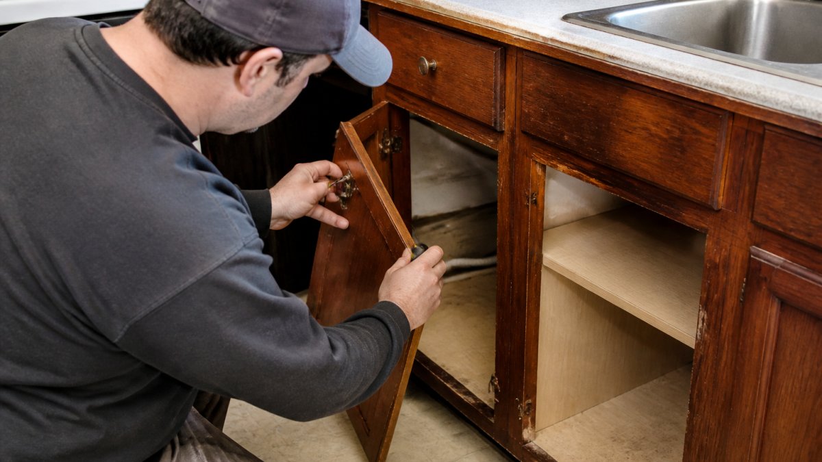 A homeowner in work clothes carefully removing a lower kitchen cabinet door with a screwdriver, exposing the base cabinet interior and shelves in an older kitchen with worn countertops