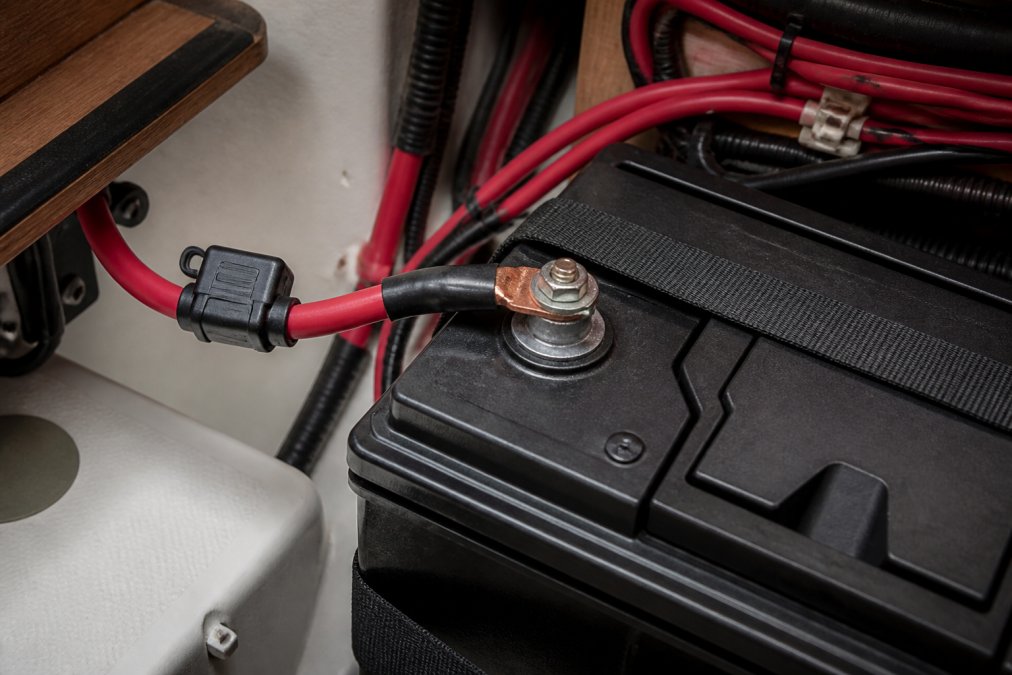 Close-up of a marine battery's positive terminal with a crimped terminal lug, inline fuse holder, and heavy-gauge tinned copper wire secured with a through-bolt and lock washer on a battery bank in a boat cabin