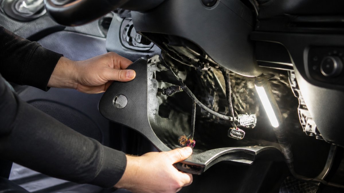 Technician's hands removing dashboard trim panels from Audi A3 8V, showing exposed connectors and wiring harness beneath steering column with work light illuminating the area