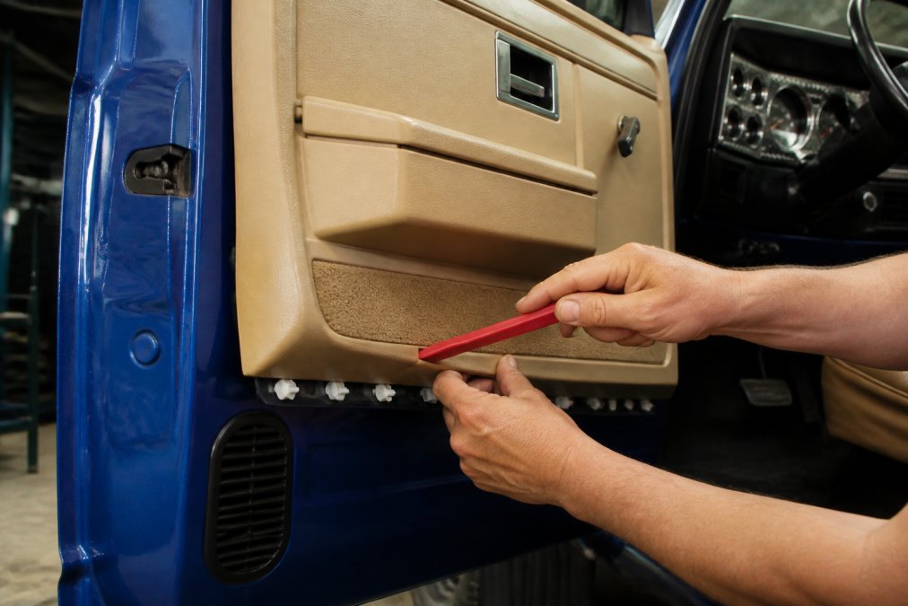 Close-up of a person's hands using a red plastic pry tool to pop a door panel clip free from the door frame of a classic OBS Chevy truck cab, with the panel partially lifted and several white plastic retaining clips visible along the lower edge, workshop lighting overhead