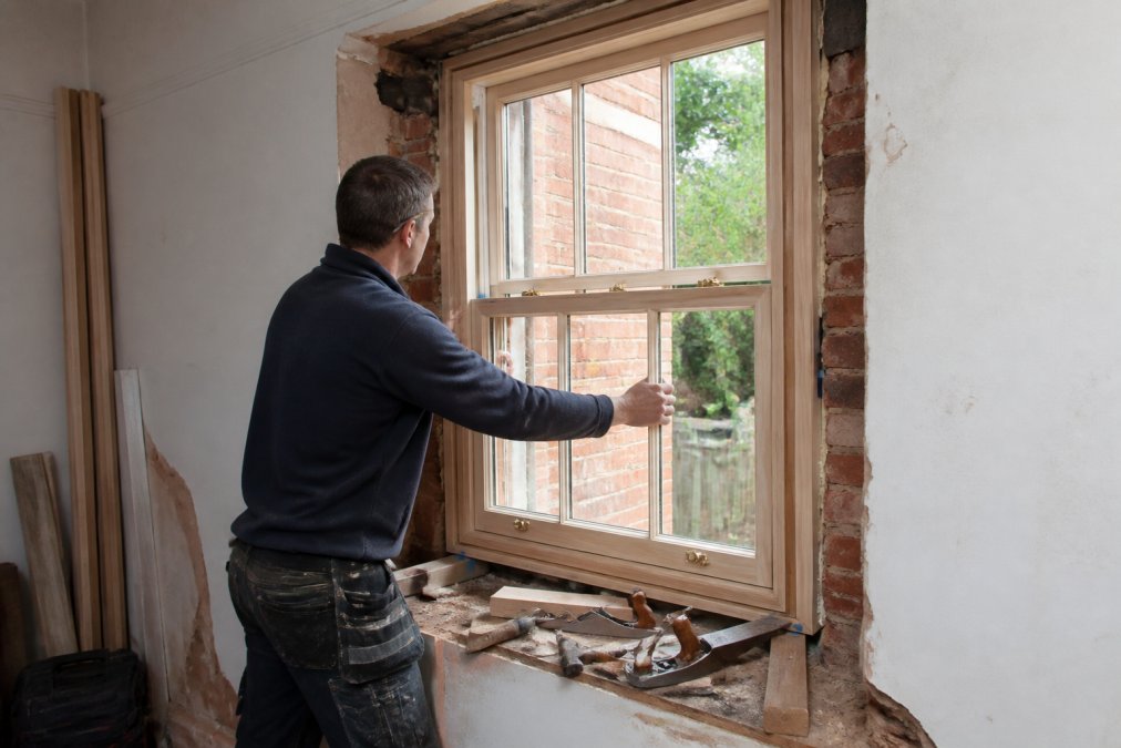 A joiner in work clothes fitting a bespoke timber sash window frame into a period property opening, with tools and timber offcuts visible nearby