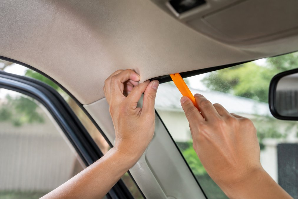 Close-up of hands using a trim removal tool to carefully peel back SUV headliner near the windshield during dash cam installation, showing proper technique to avoid cracking interior panels