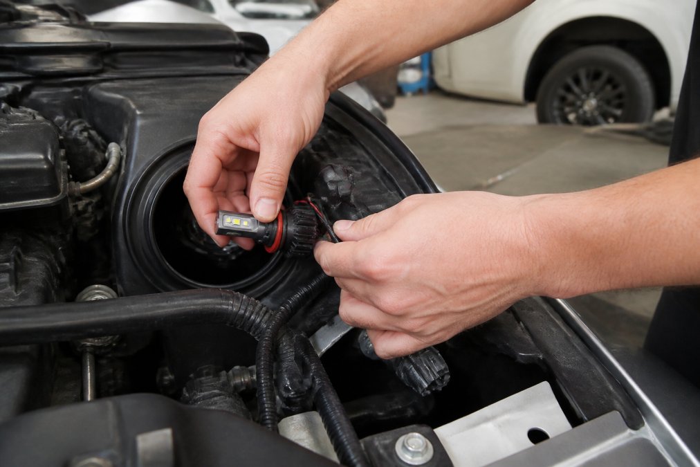 Close-up of hands installing an H11 LED bulb into a car headlight housing, with the dust cover removed and wiring harness visible, in a garage setting with natural light