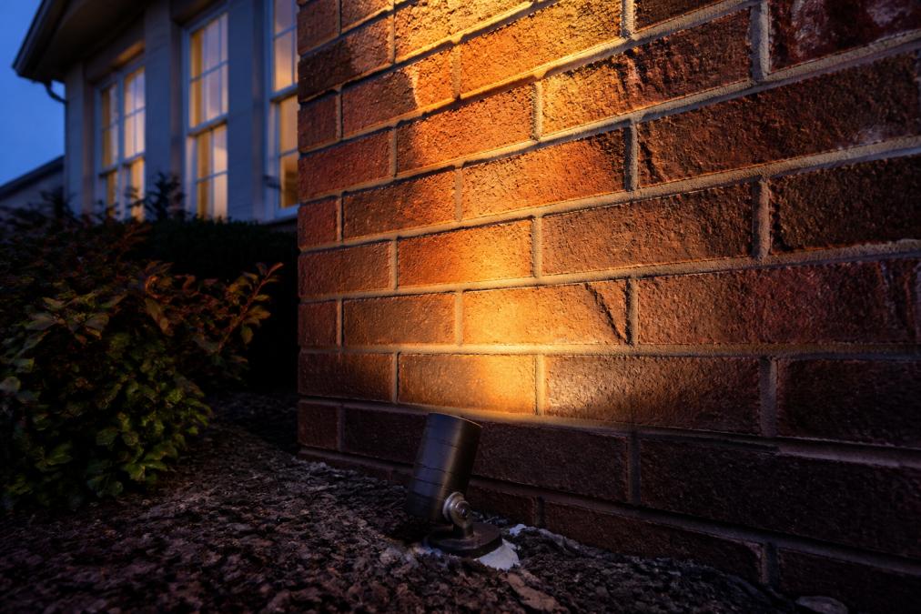Close-up of a brick facade illuminated by warm low voltage uplighting at dusk, showing fixture placement and beam spread on masonry texture, with visible caulking around fixture base