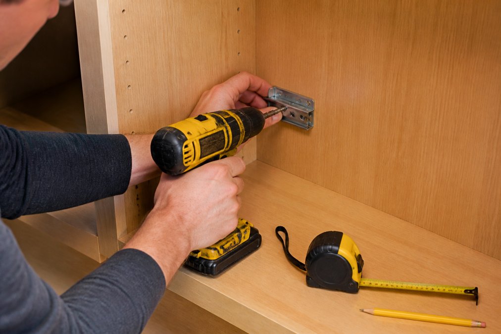 Using a power drill to attach a mounting bracket while learning how to install pull out shelves in corner cabinets