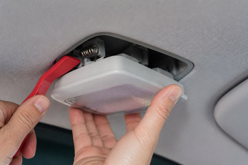 Close-up of hands carefully removing a dome light housing from a vehicle ceiling using a trim removal tool, showing the spring clip being released and the plastic housing being gently pried down