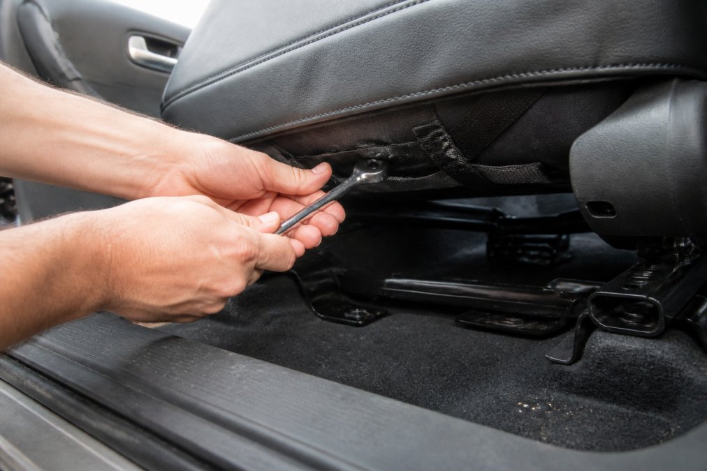 Professional hands tightening elastic straps and fasteners under a car seat with wrench, showing proper tension technique on automotive upholstery with visible seat tracks