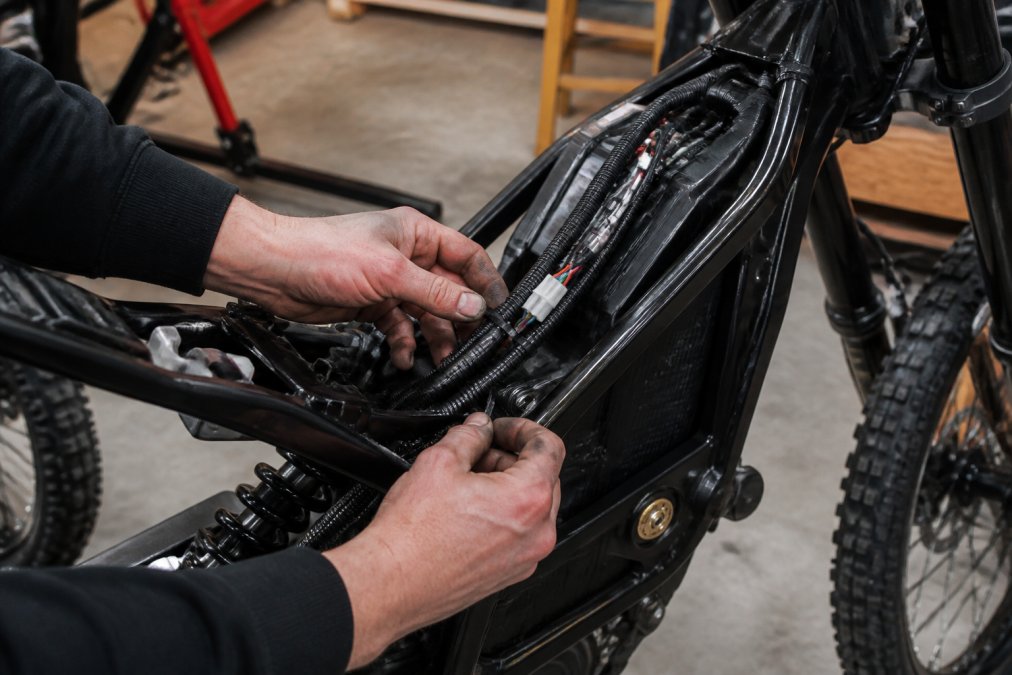Hands securing a new surron wiring harness to the electric bike frame with zip ties in a garage
