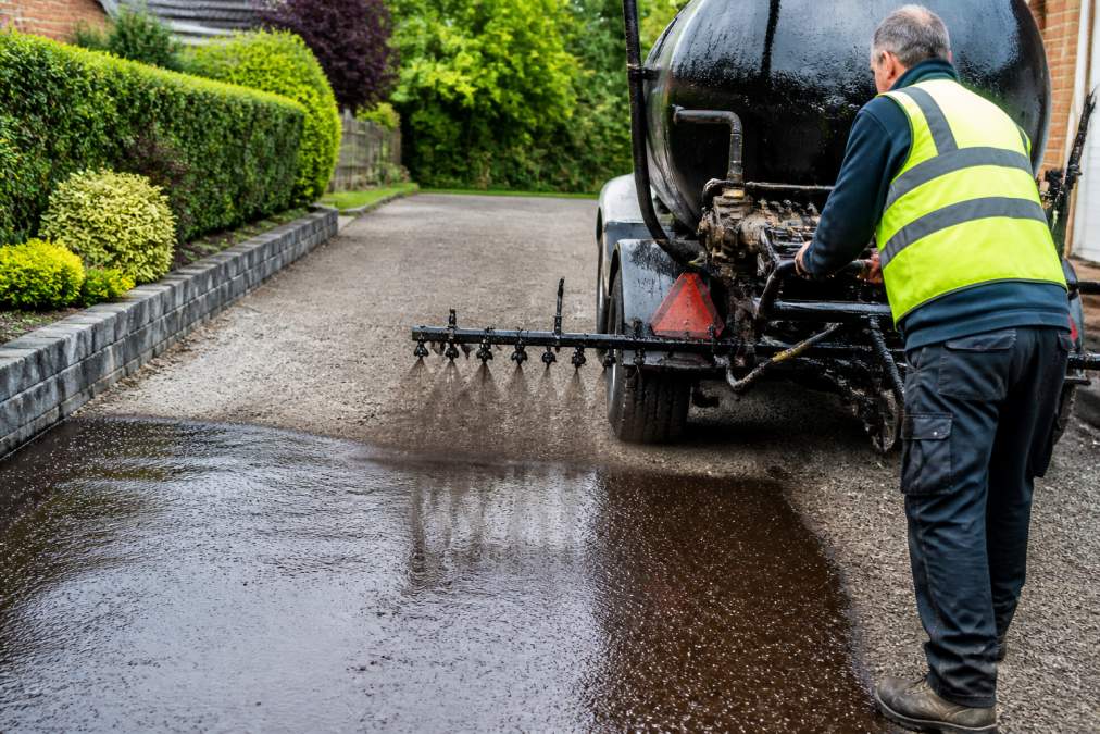 Professional paving contractor operating hot asphalt distributor equipment on residential driveway, spraying bitumen emulsion in controlled application with precision spray pattern