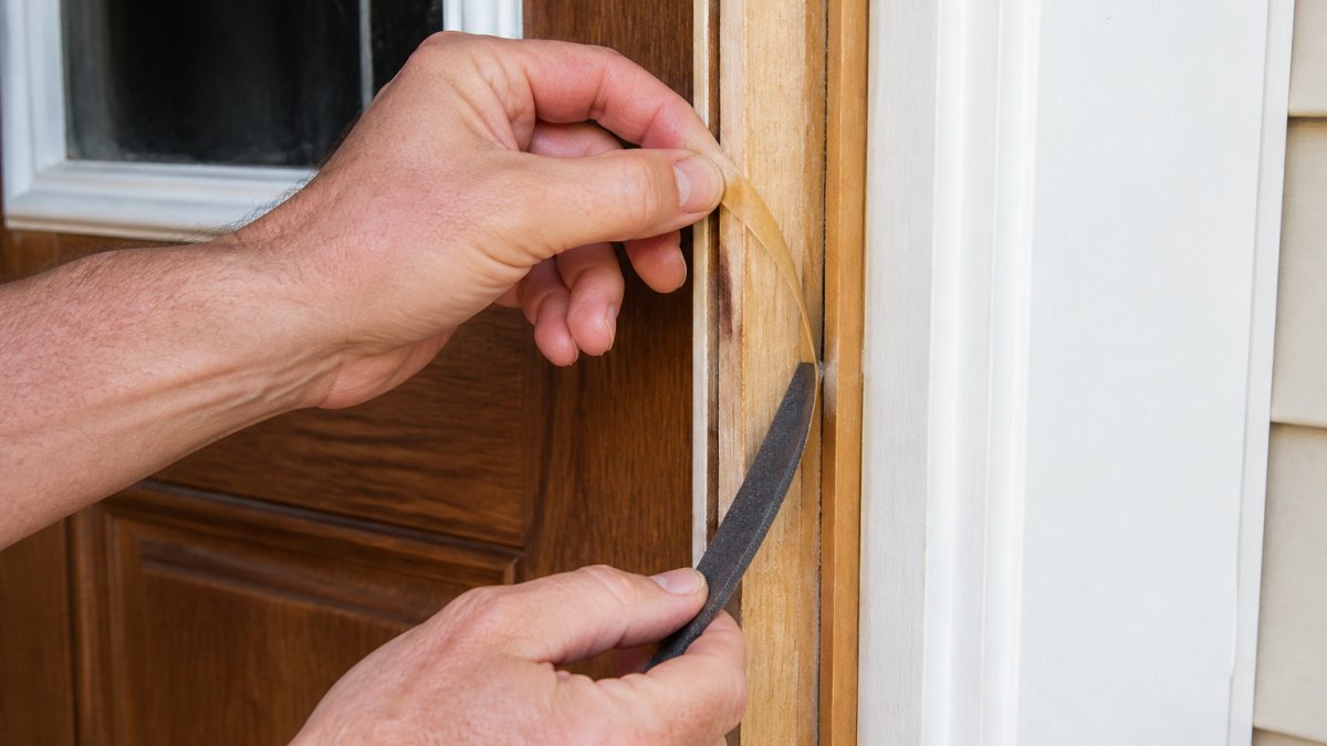 Close-up of hands carefully peeling back old adhesive-backed foam weatherstripping from a wooden door side jamb, showing the backing liner separating from the frame