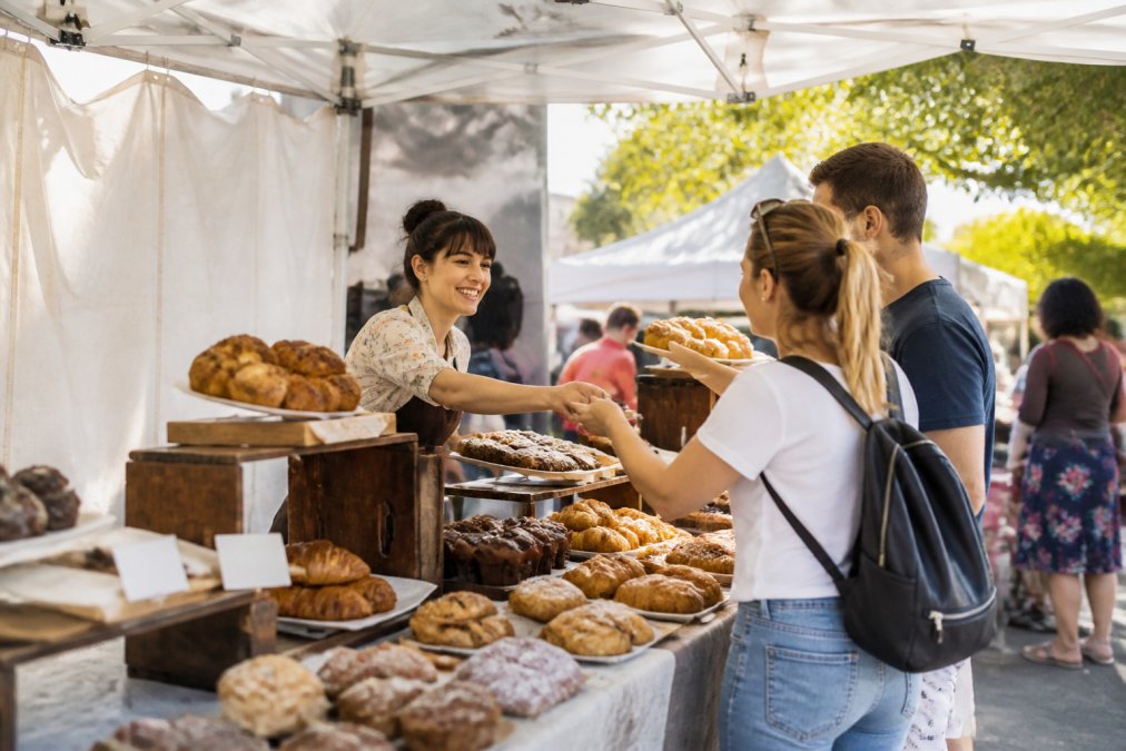 Small business owner at an outdoor farmers market booth with colorful baked goods displayed on shelves, customers browsing products, and natural sunlight illuminating the scene