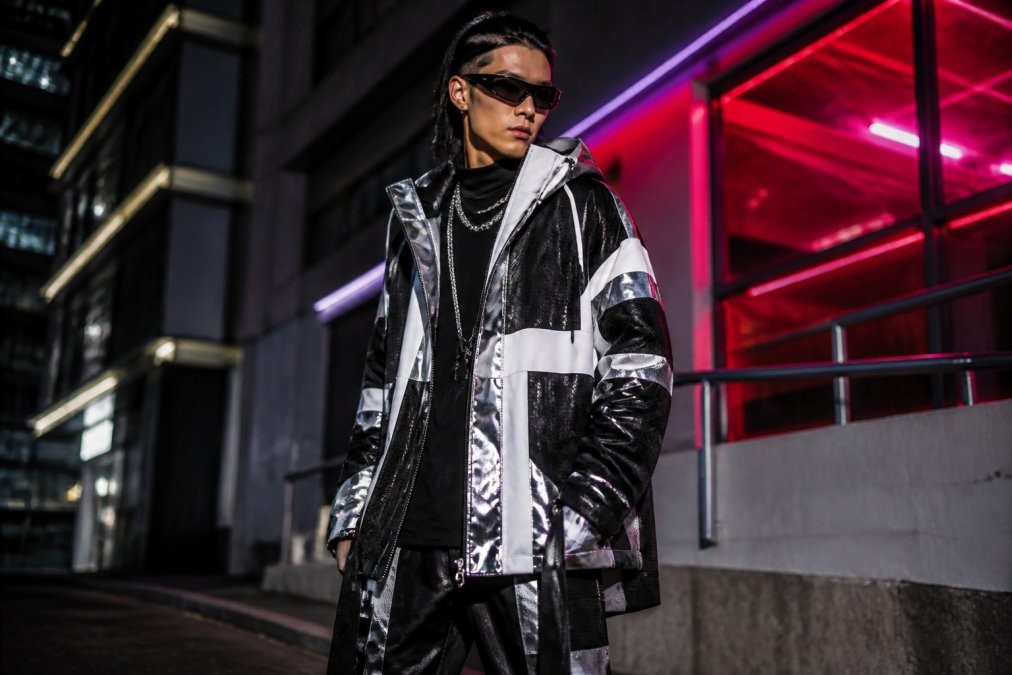 Person wearing layered futuristic streetwear with bold geometric patterns and metallic accents, posed against an urban backdrop with neon lighting