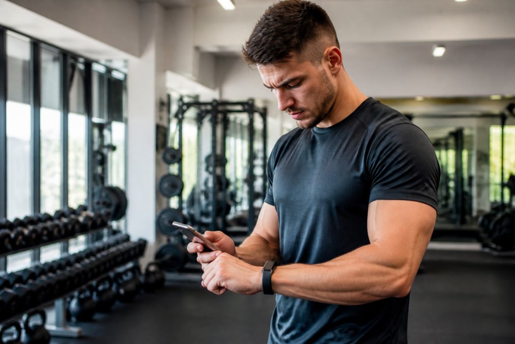 Athlete wearing a fitness tracker on their wrist while reviewing performance metrics on their smartphone in a modern gym setting with natural lighting
