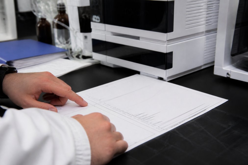 Close-up of a researcher's hands pointing at a printed LC-MS chromatogram on a lab bench, with a laptop displaying spectral data in the background