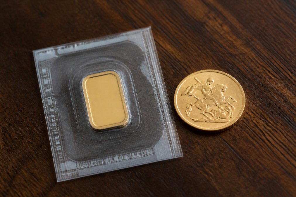 A small fine gold bar in a plastic seal beside a gold sovereign coin on a dark wooden surface with natural light