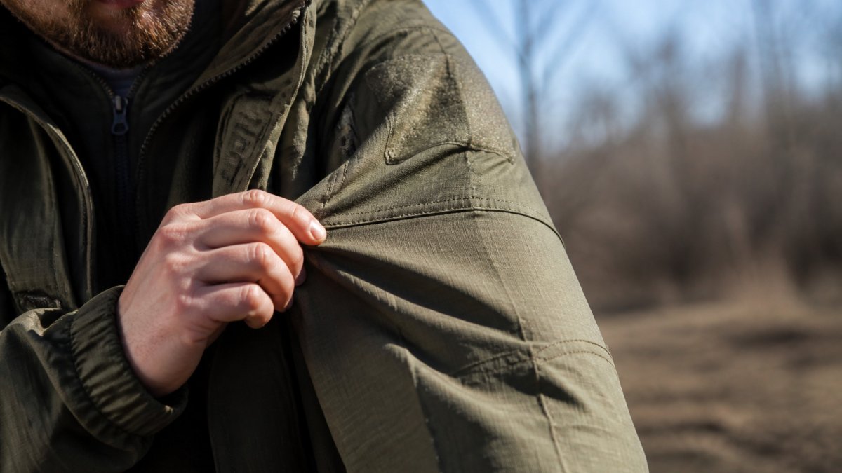 Close-up view of hands inspecting durable ripstop fabric and reinforced stitching on a field jacket outdoors