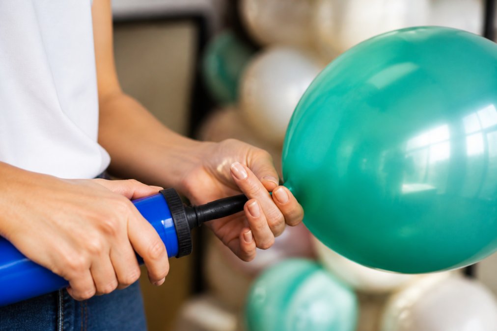 Close-up of hands properly inflating a latex balloon with a hand pump, showing correct nozzle seal technique and firm inflation pressure in bright natural lighting