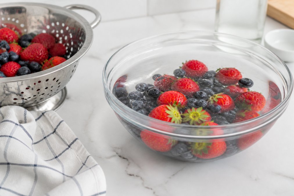 Close-up of fresh strawberries and blueberries soaking in a clear glass bowl filled with water and white vinegar on a clean white kitchen counter, with a colander nearby